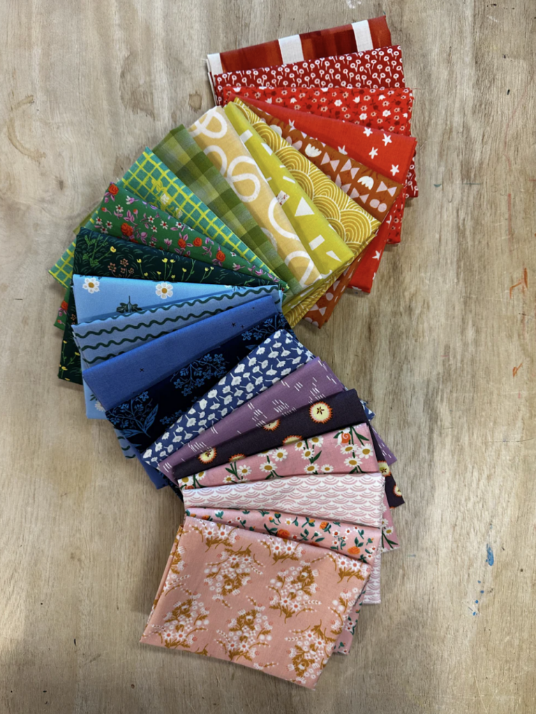 A vibrant rainbow fabric bundle arranged in a curved fan shape on a wooden surface. Fabrics range from deep red, orange, and yellow to green, blue, purple, and pink, featuring a mix of playful prints, florals, and geometric designs—perfect for a colorful version of the Front Porch Quilt Along 2025.
